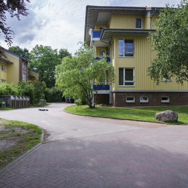 Im Hintergund: Eine gelbe Wohnunterkunft, vorne: Eine Weg und Bäume mit grünen Blättern.