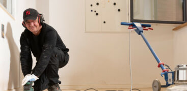 Daniel trägt Arbeitskleidung und schleift den Fußboden des Hinz&Kunzt-Hauses und lächelt in die Kamera.