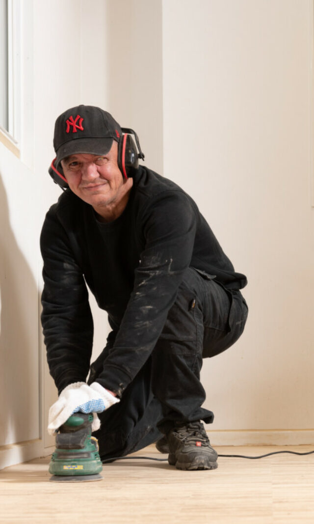 Daniel trägt Arbeitskleidung und schleift den Fußboden des Hinz&Kunzt-Hauses und lächelt in die Kamera.