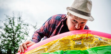 Hinz&Kunzt-Kolumnist Benjamin Buchholz trägt einen Gärtnerhut und pustet einen quietschbunten Kinderpool auf. Er schaut dabei direkt in die Kamera.