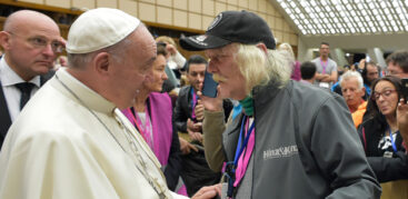 Papst Franziskus gibt einem Mann mit schulterlangen weißen Haaren und einer schwarzen Mütze die Hand