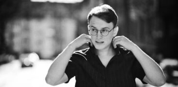 Schwarz-weiß-Foto von einem jungen Mann mit runder Brille im schwarzen Hemd