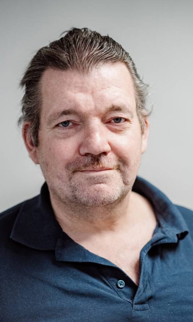 Ein mittelalter Mann im dunkelblauen Polohemd schaut mit festem Blick in die Kamera