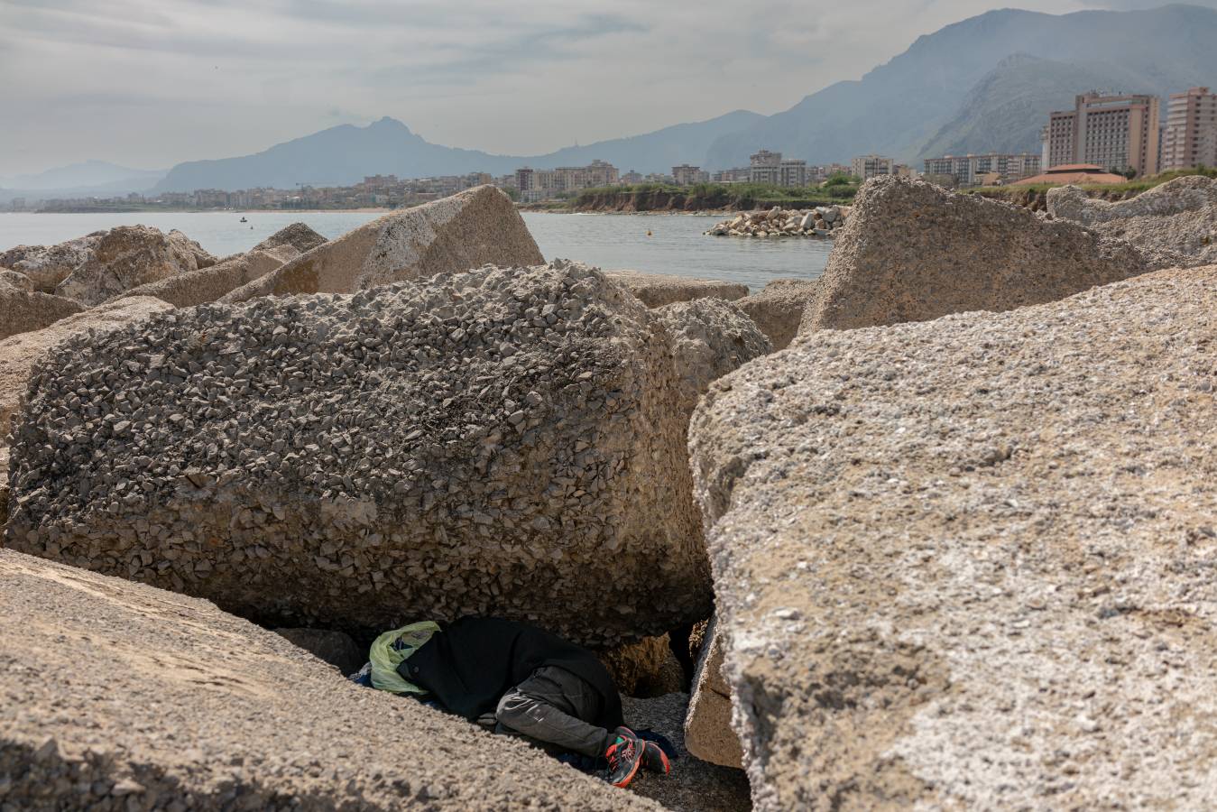 Große Steine an einer Küste, dazwischen liegt zusammengekauert ein Mensch