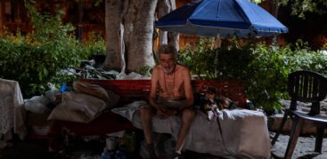 Ein Mann mit nacktem Oberkörper sitzt mit einem Hund auf einer Decke, die auf einer Bank liegt