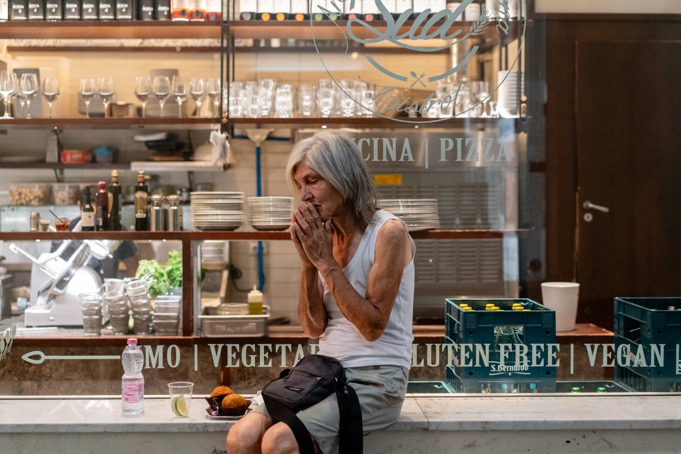 Eine grauhaarige hagere Frau sitzt mit etwas Gebäck vor dem Schaufenster einer Pizzeria