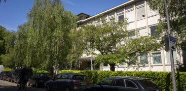 Ein dreistöckiges Haus hinter Bäumen, an der Straße davor stehen Autos