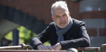 Ein Mann mit schwarzem Sakko und grauem Schal lehnt auf einem Geländer