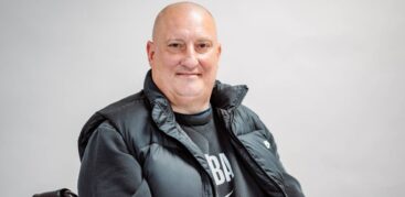 Ein glatzköpfiger Mann sitzt auf seinem Rollator und lacht freundlich in die Kamera