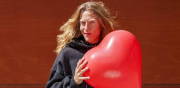 Eine Frau mit roten Haaren und Stachelhalsband hält einen herzförmigen Luftballon.