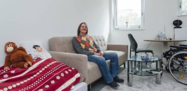 Ein Mann im karierten Pullover sitzt in einem hellen Zimmer auf einem Sofa