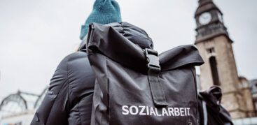 Ein Mensch mit grüner Mütze von hinten, auf dem Rucksack steht "Sozialarbeit"