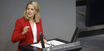 Eine blonde Frau im roten Sakko steht am Redner:innenpult des Bundestags.