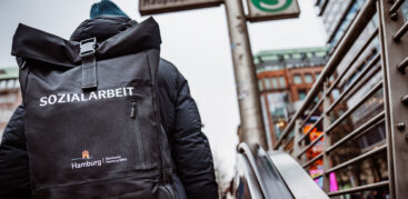Eine Frau ist von hinten zu sehen, wie sie Rolltreppen am Hauptbahnhof hochfährt. Auf ihrem Rucksack steht "Straßensozialarbeit".
