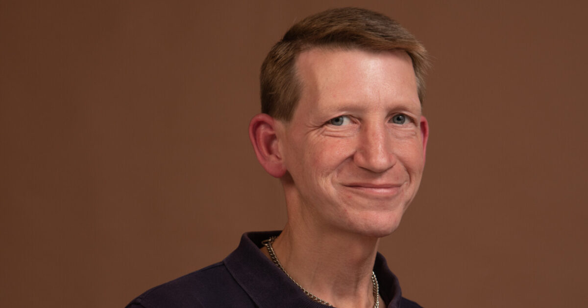 Ein blonder Mann im Polohemd lacht in die Kamera