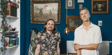 Die beiden Autoren Nikolas Kuhl und Stefan Sandrock stehen vor einer blauen Wand mit zahlreichen Bildrahmen. Foto: Miguel Ferraz