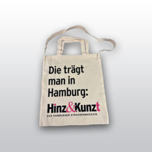 Die Abbildung zeigt eine helle Baumwolltasche mit Henkeln und einem langen Trageriemen. Auf der Tasche ist in schwarzer Schrift zu lesen: "Die trägt man in Hamburg: Hinz&Kunzt DAS HAMBURGER STRASSENMAGAZIN".