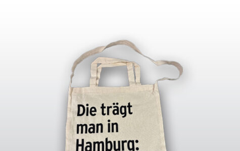 Die Abbildung zeigt eine helle Baumwolltasche mit Henkeln und einem langen Trageriemen. Auf der Tasche ist in schwarzer Schrift zu lesen: "Die trägt man in Hamburg: Hinz&Kunzt DAS HAMBURGER STRASSENMAGAZIN".