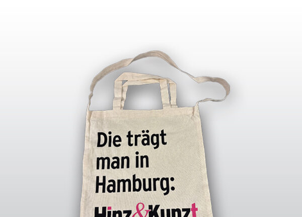 Die Abbildung zeigt eine helle Baumwolltasche mit Henkeln und einem langen Trageriemen. Auf der Tasche ist in schwarzer Schrift zu lesen: "Die trägt man in Hamburg: Hinz&Kunzt DAS HAMBURGER STRASSENMAGAZIN".