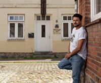 Ein Mann im weißen T-Shirt lehnt an einer Hauswand, im Hintergrund ein beiges Haus