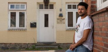 Ein Mann im weißen T-Shirt lehnt an einer Hauswand, im Hintergrund ein beiges Haus