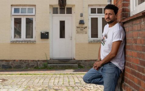 Ein Mann im weißen T-Shirt lehnt an einer Hauswand, im Hintergrund ein beiges Haus