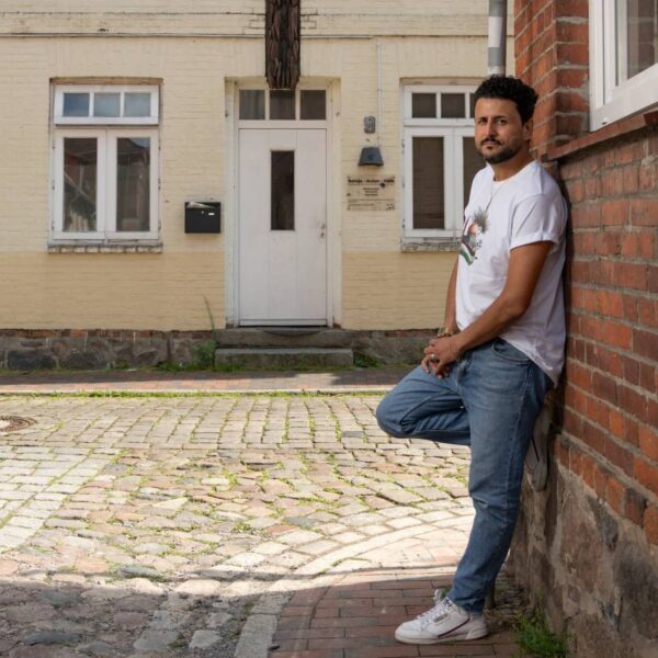 Ein Mann im weißen T-Shirt lehnt an einer Hauswand, im Hintergrund ein beiges Haus