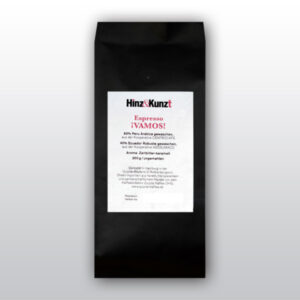 Das Bild zeigt eine schwarze Kaffeeverpackung mit einem weißen Etikett in der Mitte. Auf dem Etikett steht in roter und schwarzer Schrift "Hinz & Kunzt Espresso ¡VAMOS!". Darunter sind Details zum Kaffee aufgeführt