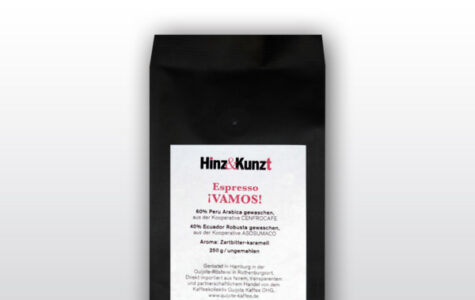 Das Bild zeigt eine schwarze Kaffeeverpackung mit einem weißen Etikett in der Mitte. Auf dem Etikett steht in roter und schwarzer Schrift "Hinz & Kunzt Espresso ¡VAMOS!". Darunter sind Details zum Kaffee aufgeführt