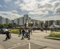 Fußgänger:innen gehen über eine Ampel, im Hintergrund eine gewaltige Reihe von Hochhäusern