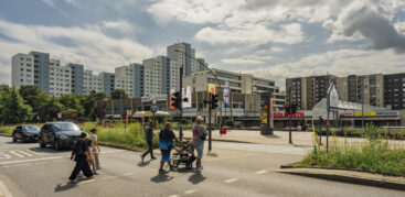 Fußgänger:innen gehen über eine Ampel, im Hintergrund eine gewaltige Reihe von Hochhäusern