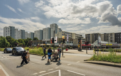 Fußgänger:innen gehen über eine Ampel, im Hintergrund eine gewaltige Reihe von Hochhäusern