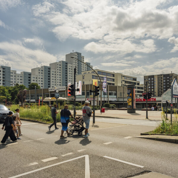 Fußgänger:innen gehen über eine Ampel, im Hintergrund eine gewaltige Reihe von Hochhäusern