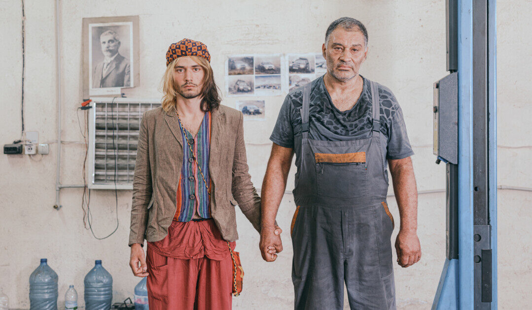 Zwei Männer halten Händchen in einer Autowerkstatt und schauen ausdruckslos in die Kamera