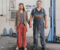 Zwei Männer halten Händchen in einer Autowerkstatt und schauen ausdruckslos in die Kamera