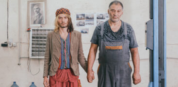 Zwei Männer halten Händchen in einer Autowerkstatt und schauen ausdruckslos in die Kamera