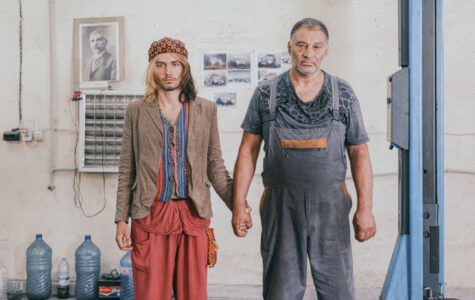 Zwei Männer halten Händchen in einer Autowerkstatt und schauen ausdruckslos in die Kamera