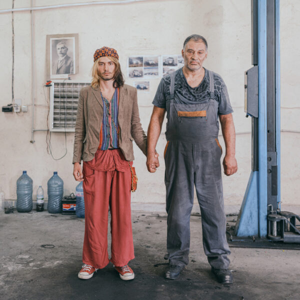 Zwei Männer halten Händchen in einer Autowerkstatt und schauen ausdruckslos in die Kamera