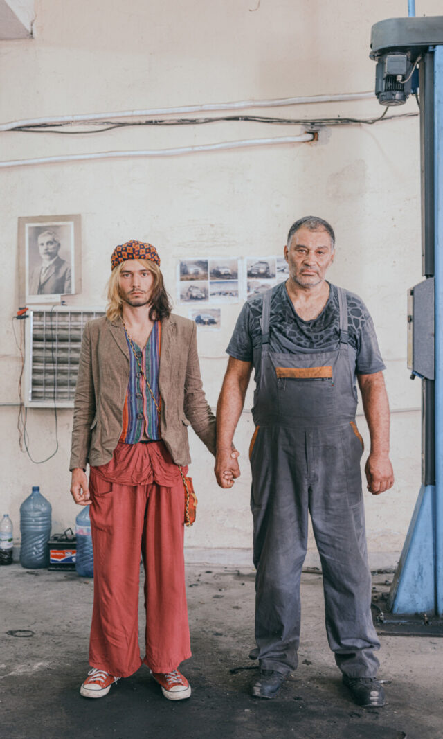 Zwei Männer halten Händchen in einer Autowerkstatt und schauen ausdruckslos in die Kamera