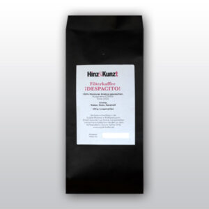 Das Bild zeigt eine schwarze Kaffeeverpackung mit einem weißen Etikett in der Mitte. Auf dem Etikett steht in roter und schwarzer Schrift "Hinz & Kunzt Espresso ¡DESOACITO!". Darunter sind Details zum Kaffee aufgeführt