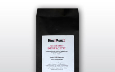 Das Bild zeigt eine schwarze Kaffeeverpackung mit einem weißen Etikett in der Mitte. Auf dem Etikett steht in roter und schwarzer Schrift "Hinz & Kunzt Espresso ¡DESOACITO!". Darunter sind Details zum Kaffee aufgeführt