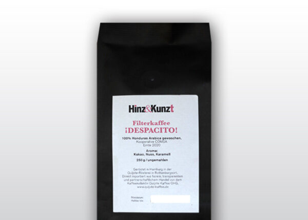 Das Bild zeigt eine schwarze Kaffeeverpackung mit einem weißen Etikett in der Mitte. Auf dem Etikett steht in roter und schwarzer Schrift "Hinz & Kunzt Espresso ¡DESOACITO!". Darunter sind Details zum Kaffee aufgeführt