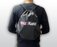 Ein schwarzer Turnbeutel mit Kordelzug ist auf dem Rücken einer Person zu sehen. Auf dem Beutel steht in weißer und pinker Schrift „Hinz & Kunz!“ und darunter in kleinerer pinker Schrift „30 JAHRE“.