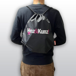 Ein schwarzer Turnbeutel mit Kordelzug ist auf dem Rücken einer Person zu sehen. Auf dem Beutel steht in weißer und pinker Schrift „Hinz & Kunz!“ und darunter in kleinerer pinker Schrift „30 JAHRE“.