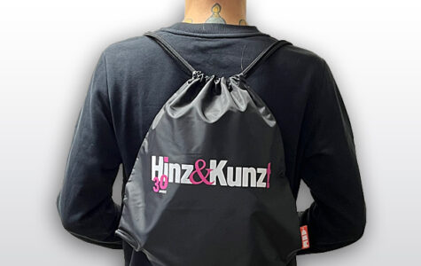 Ein schwarzer Turnbeutel mit Kordelzug ist auf dem Rücken einer Person zu sehen. Auf dem Beutel steht in weißer und pinker Schrift „Hinz & Kunz!“ und darunter in kleinerer pinker Schrift „30 JAHRE“.