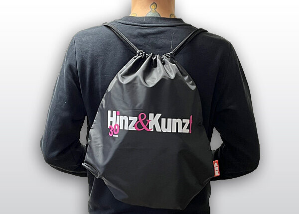 Ein schwarzer Turnbeutel mit Kordelzug ist auf dem Rücken einer Person zu sehen. Auf dem Beutel steht in weißer und pinker Schrift „Hinz & Kunz!“ und darunter in kleinerer pinker Schrift „30 JAHRE“.