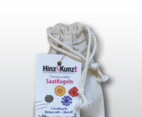 Das Bild zeigt einen naturfarbenen Stoffbeutel mit Kordelzug, der Saatkugeln enthält. An dem Beutel hängt ein weißes Etikett der Hamburger Straßenzeitung "Hinz&Kunzt". Auf dem Etikett steht prominent "Bienenweide SaatKugeln" und darunter "5 Saatkugeln Blühen soll's – überall!". Das Etikett zeigt zudem eine Illustration von fünf Saatkugeln und stilisierten Blumen in Gelb, Rot und Blau, die auf die Vielfalt der enthaltenen Samen hinweisen. Im unteren Bereich des Etiketts sind schematische Darstellungen von Häusern zu sehen