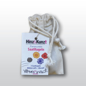 Das Bild zeigt einen naturfarbenen Stoffbeutel mit Kordelzug, der Saatkugeln enthält. An dem Beutel hängt ein weißes Etikett der Hamburger Straßenzeitung "Hinz&Kunzt". Auf dem Etikett steht prominent "Bienenweide SaatKugeln" und darunter "5 Saatkugeln Blühen soll's – überall!". Das Etikett zeigt zudem eine Illustration von fünf Saatkugeln und stilisierten Blumen in Gelb, Rot und Blau, die auf die Vielfalt der enthaltenen Samen hinweisen. Im unteren Bereich des Etiketts sind schematische Darstellungen von Häusern zu sehen