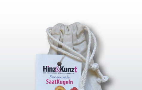 Das Bild zeigt einen naturfarbenen Stoffbeutel mit Kordelzug, der Saatkugeln enthält. An dem Beutel hängt ein weißes Etikett der Hamburger Straßenzeitung "Hinz&Kunzt". Auf dem Etikett steht prominent "Bienenweide SaatKugeln" und darunter "5 Saatkugeln Blühen soll's – überall!". Das Etikett zeigt zudem eine Illustration von fünf Saatkugeln und stilisierten Blumen in Gelb, Rot und Blau, die auf die Vielfalt der enthaltenen Samen hinweisen. Im unteren Bereich des Etiketts sind schematische Darstellungen von Häusern zu sehen