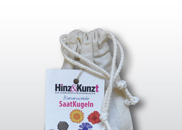 Das Bild zeigt einen naturfarbenen Stoffbeutel mit Kordelzug, der Saatkugeln enthält. An dem Beutel hängt ein weißes Etikett der Hamburger Straßenzeitung "Hinz&Kunzt". Auf dem Etikett steht prominent "Bienenweide SaatKugeln" und darunter "5 Saatkugeln Blühen soll's – überall!". Das Etikett zeigt zudem eine Illustration von fünf Saatkugeln und stilisierten Blumen in Gelb, Rot und Blau, die auf die Vielfalt der enthaltenen Samen hinweisen. Im unteren Bereich des Etiketts sind schematische Darstellungen von Häusern zu sehen